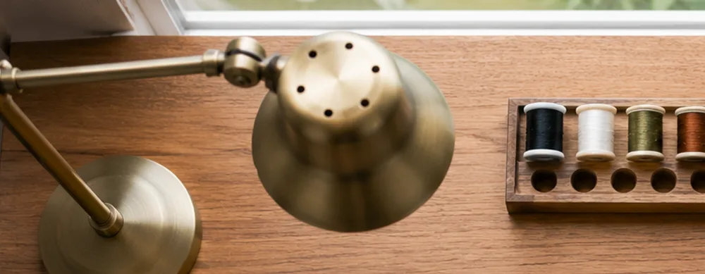 Brass desk lamp angled over a wooden bench beside a rack of thread spools, daylight from a window